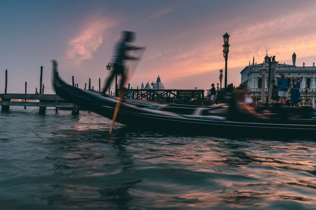 A dynamic evening scene in Venice, featuring a gondolier in motion, propelling a gondola through the water at sunset. The sky is painted with warm hues of pink and orange, reflecting off the rippling water. In the background, the silhouette of a historic building and the domes of Santa Maria della Salute are visible, along with a ‘Servizio Gondole’ sign. The scene captures the romance and energy of a gondola ride during the golden hour in Venice.