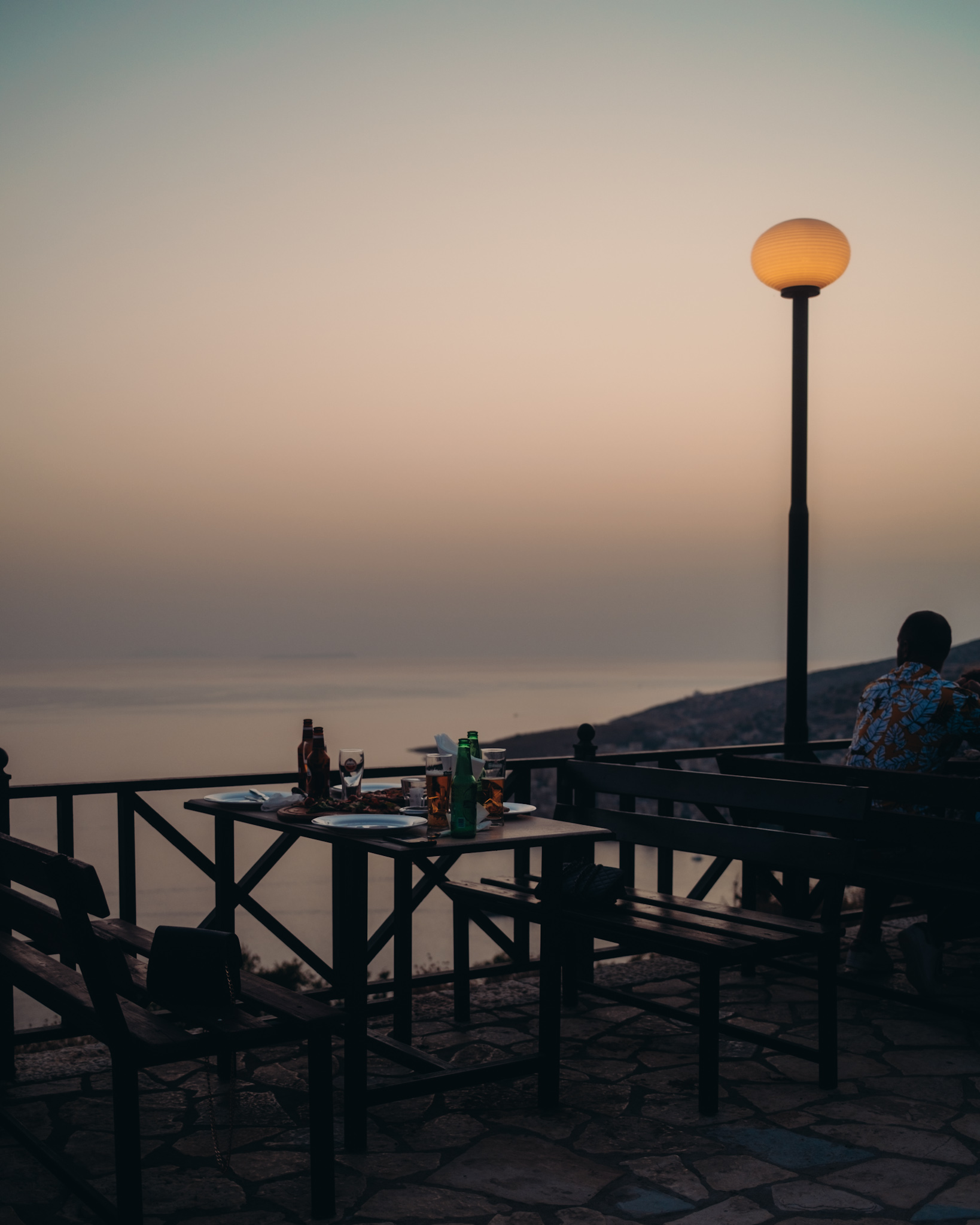 Gedeckter Restauranttisch mit Getränken auf der Terrasse des Lëkurësi Schlosses, eine Person blickt auf das Meer bei Sonnenuntergang.