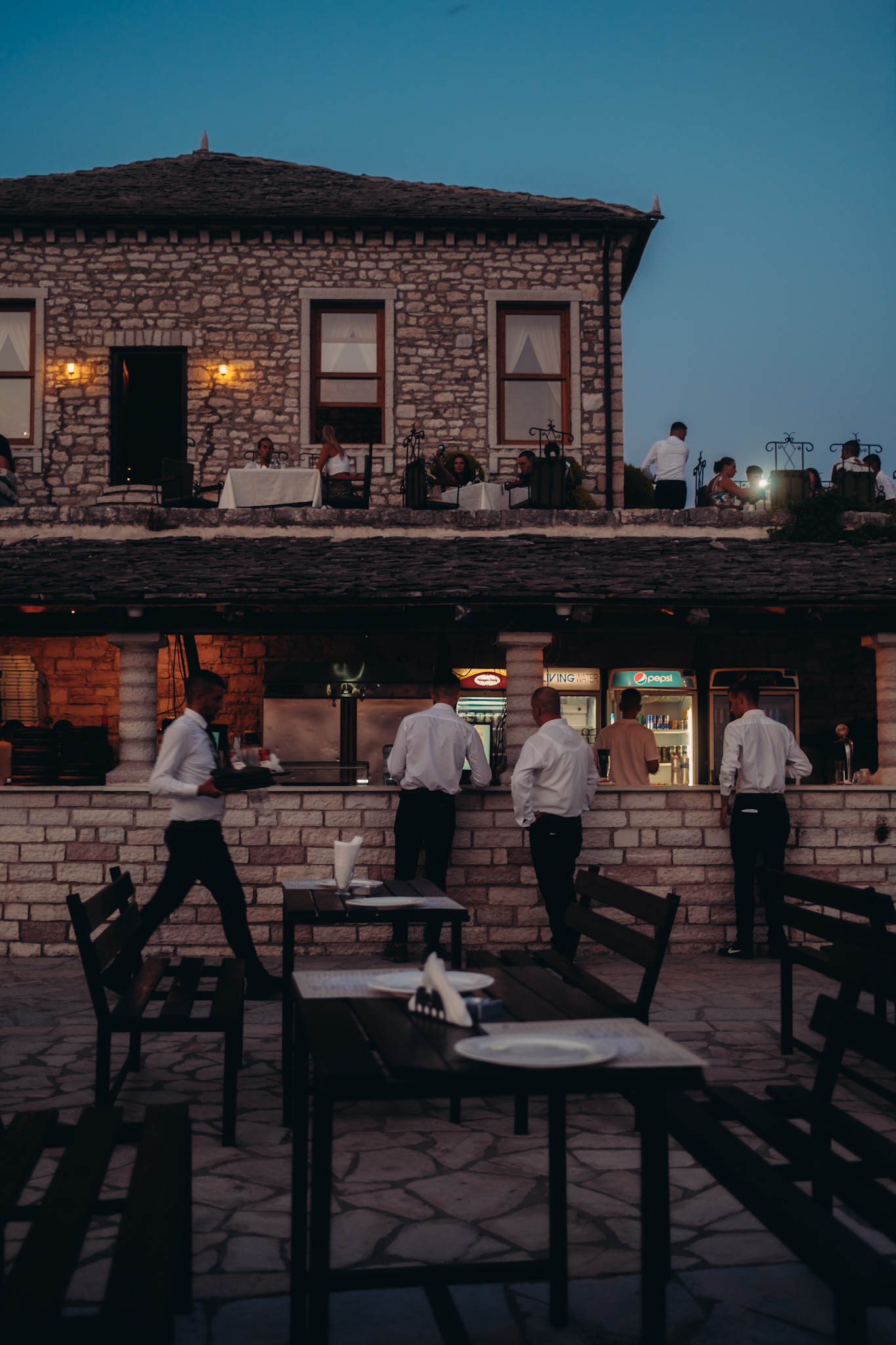 Steingebäude des Restaurants im Lëkurësi Schloss in Saranda bei Nacht, mit beleuchteten Fenstern, Kellnern und Gästen auf der Terrasse.