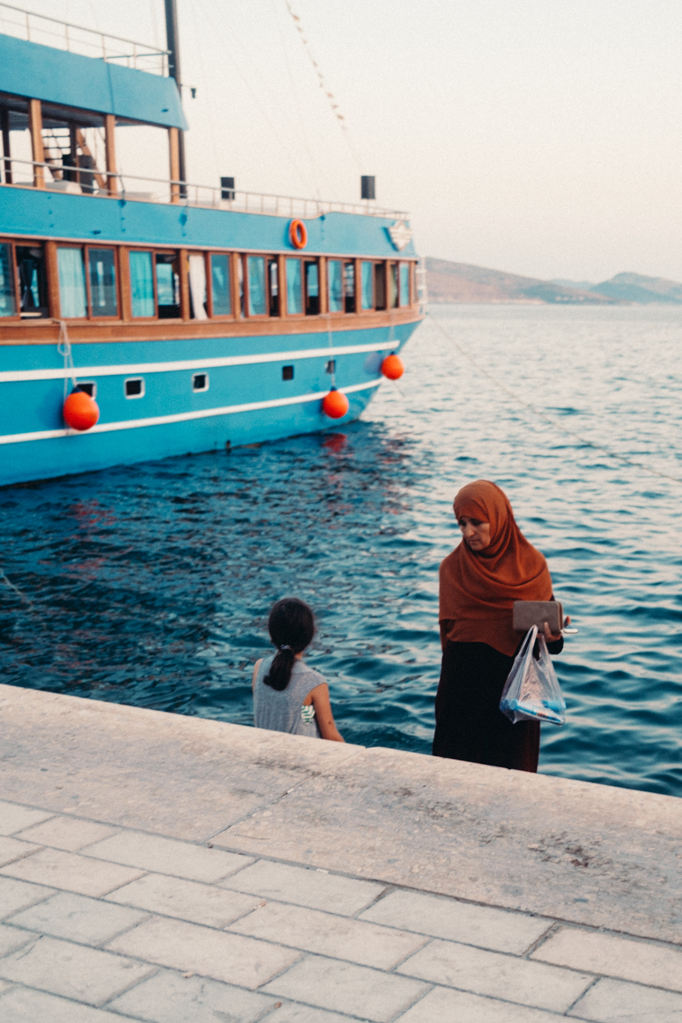 Eine Frau mit Kopftuch und ein Kind stehen am Kai in Saranda neben einem großen blauen Schiff.