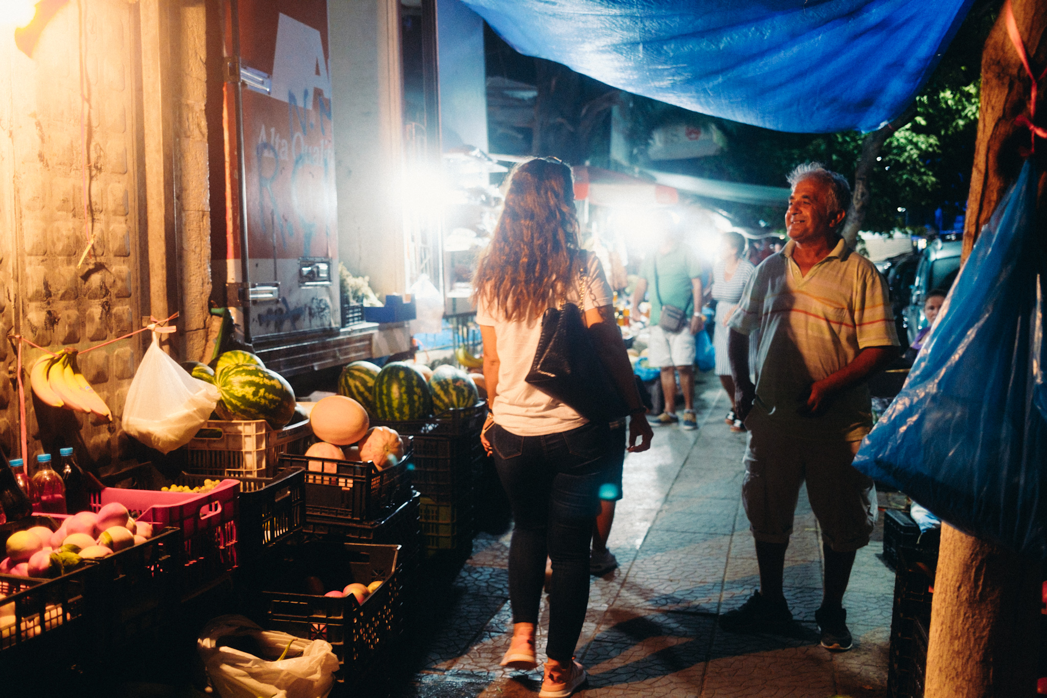 Nächtliche Marktszene in Saranda: Eine Frau betrachtet Obst an einem Stand, ein Mann steht daneben, beleuchtet von Lampen.