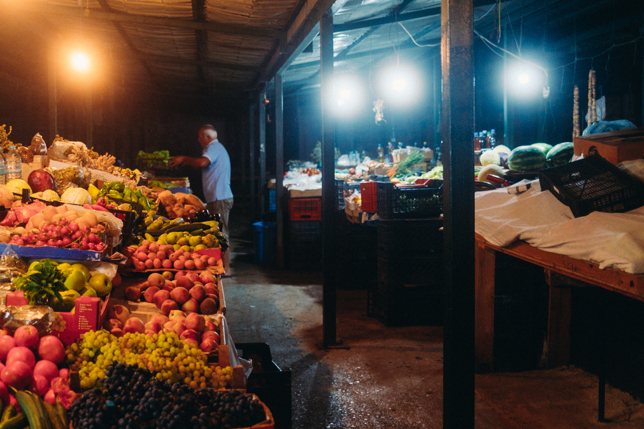 Ein üppig bestückter Obst- und Gemüsestand auf einem Markt in Saranda bei Nacht, ein Mann steht im Hintergrund.