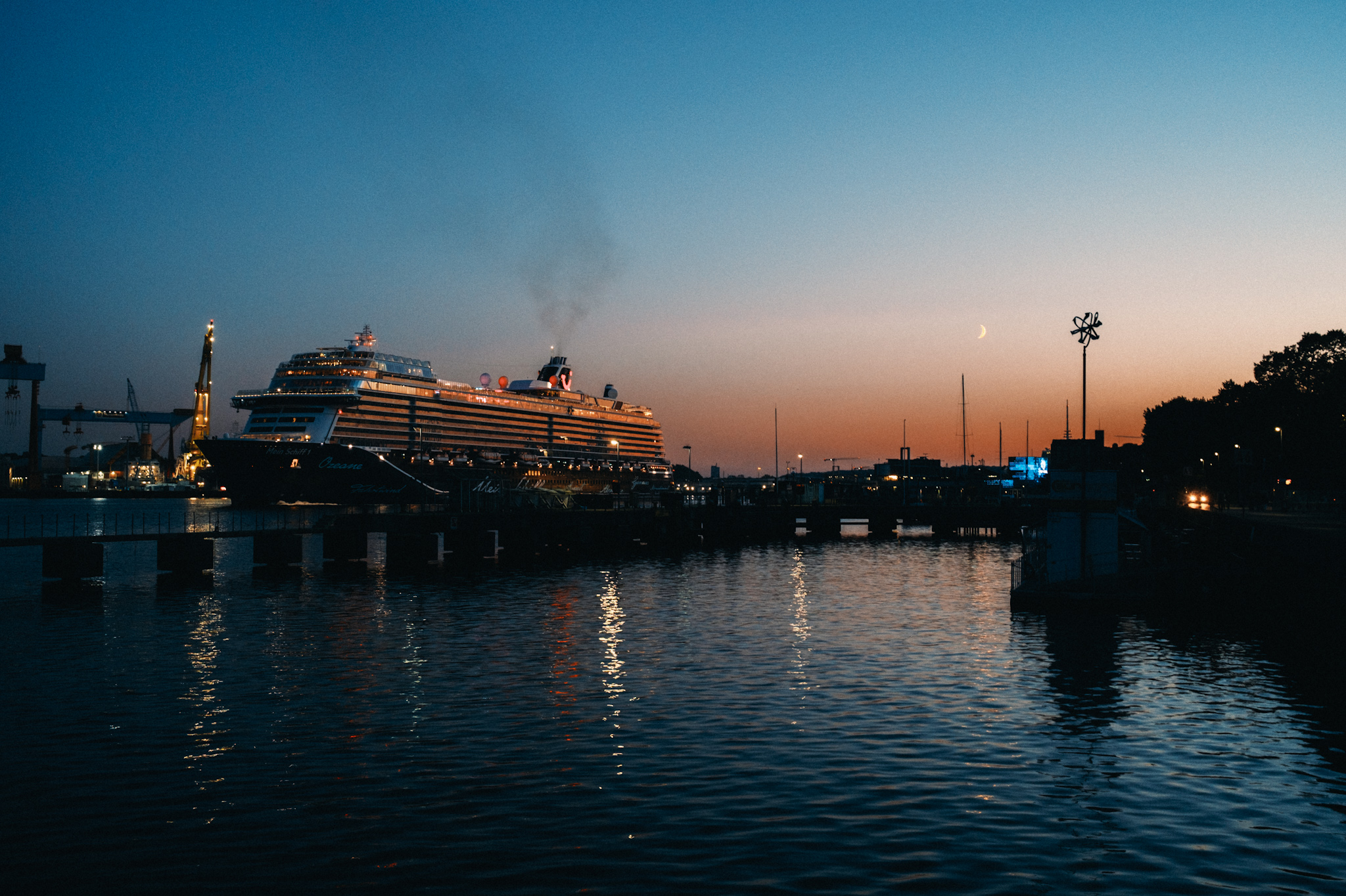 Das Kreuzfahrtschiff "Mein Schiff" aus mittlerer Distanz an der Kiellinie in Kiel, mit Hafenkulisse, einer Mondsichel am dämmernden Himmel und Spiegelungen im Wasser.