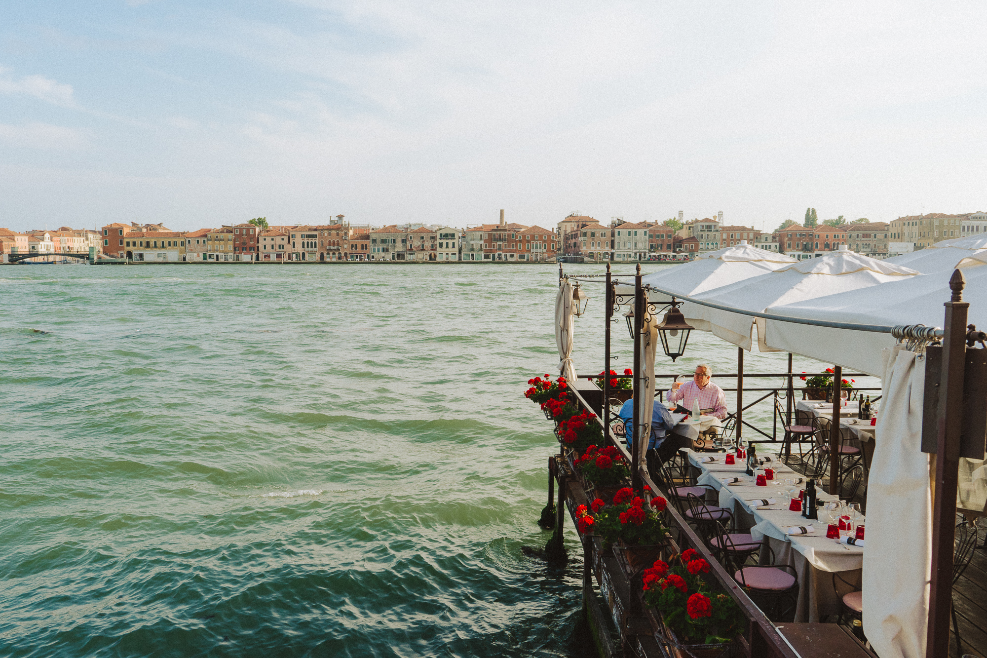 A picturesque outdoor dining area set on a wooden deck along the Venetian waterfront, adorned with vibrant red flowers. Two men are seated at a table under white canopies, enjoying a meal with a view of the calm green waters. In the background, the island of Giudecca is visible, lined with pastel-colored historic buildings under a bright, clear sky.