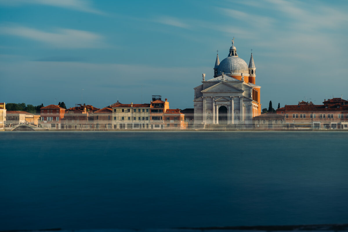 A long exposure shot of the Church of the Most Holy Redeemer (Il Redentore) in Venice during sunset. The image captures the stately church with its domes and classical facade, bathed in the warm light of the setting sun. A ghostly streak, likely from a passing boat, blurs across the water in the foreground, adding a dynamic element to the tranquil scene. The background features the soft pastel hues of the surrounding historic buildings against a clear sky.