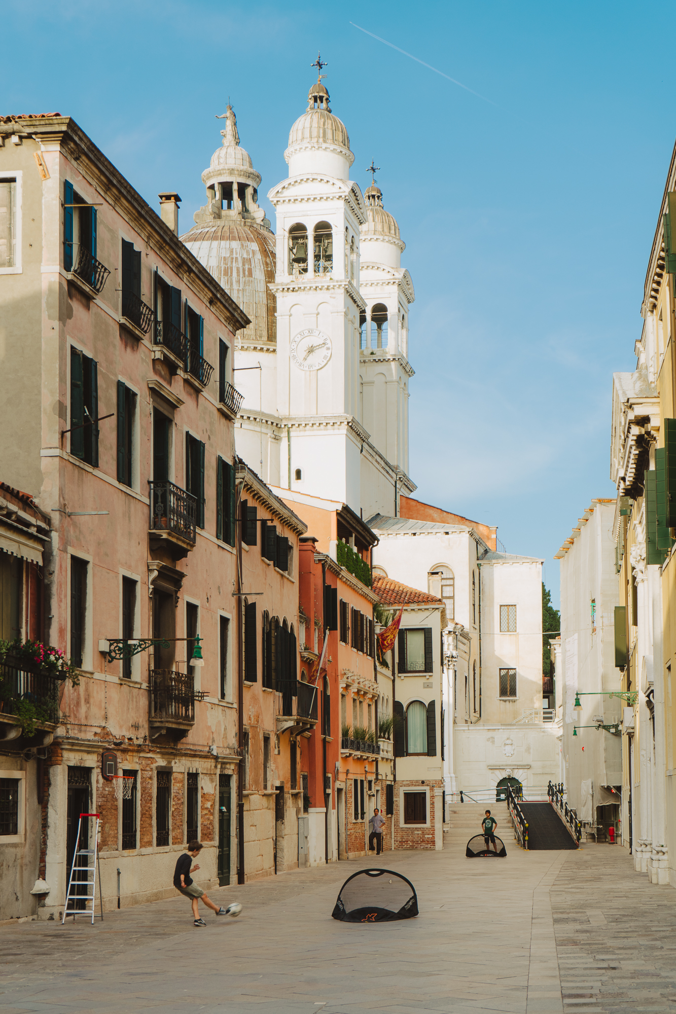 Two children playing an energetic game of street soccer on a narrow Venetian street, surrounded by colorful historic buildings. In the background, the towering white domes and bell tower of the Church of Santi Giovanni e Paolo rise against a clear blue sky, adding a dramatic architectural contrast to the lively scene.