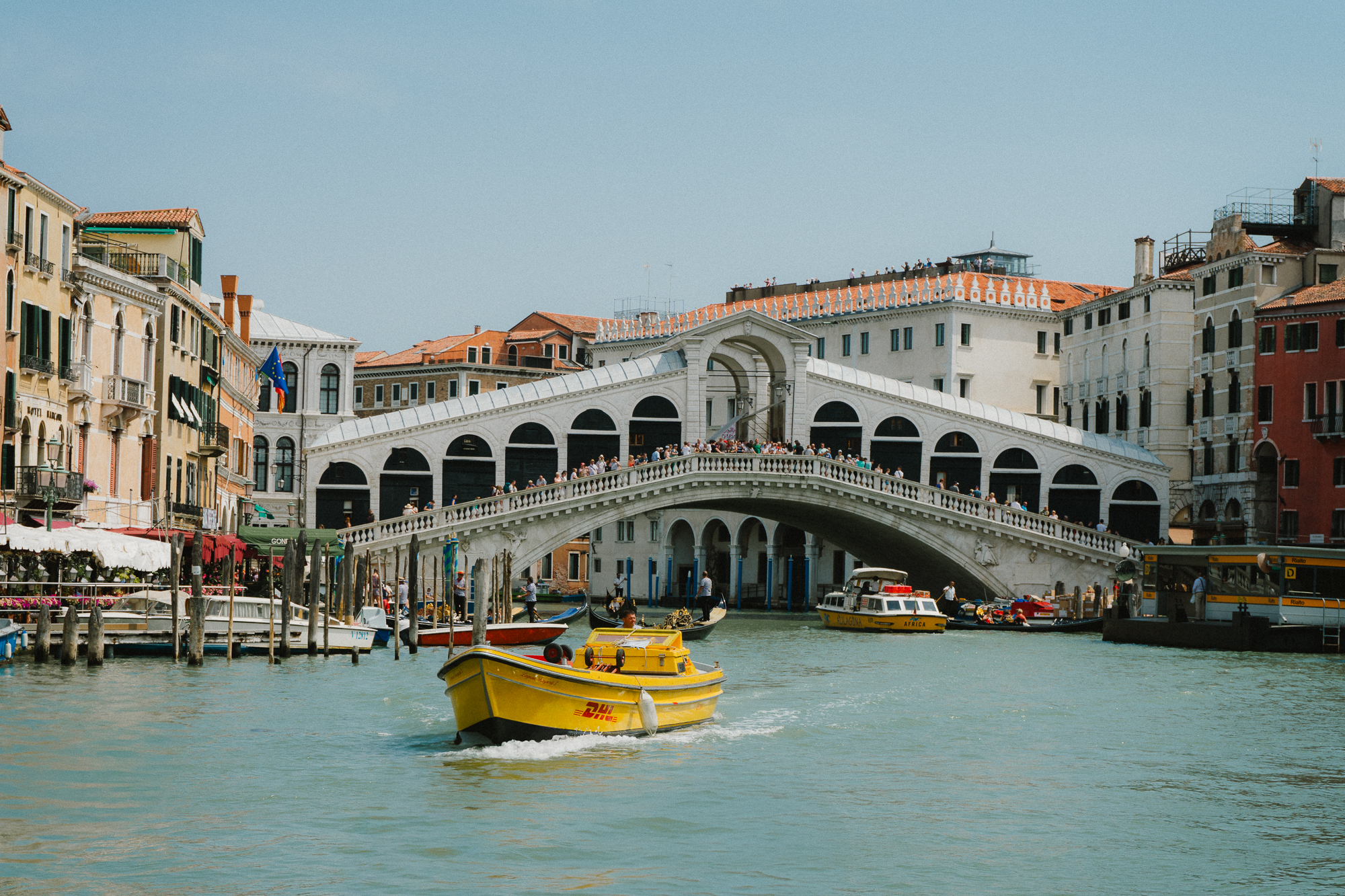 A vibrant scene on Venice’s Grand Canal, featuring the famous Rialto Bridge. The bridge, bustling with tourists, arches gracefully over the canal, connecting the historic buildings on either side. In the foreground, a bright yellow water taxi navigates the canal, surrounded by other boats and gondolas, all adding to the lively atmosphere of the Venetian waterway. The buildings lining the canal display typical Venetian architecture with colorful facades under a clear blue sky.