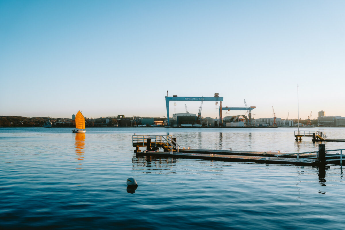 A striking golden sail glides across the Kiel Fjord, set against the industrial backdrop of German Naval Yards. A harmonious blend of tradition and modernity along the Kiellinie.