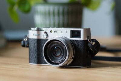 Fuji X100V Kamera auf einem Holztisch mit unscharfer grüner Pflanze im Hintergrund, natürliches Licht betont das Metall- und Ledergehäuse.