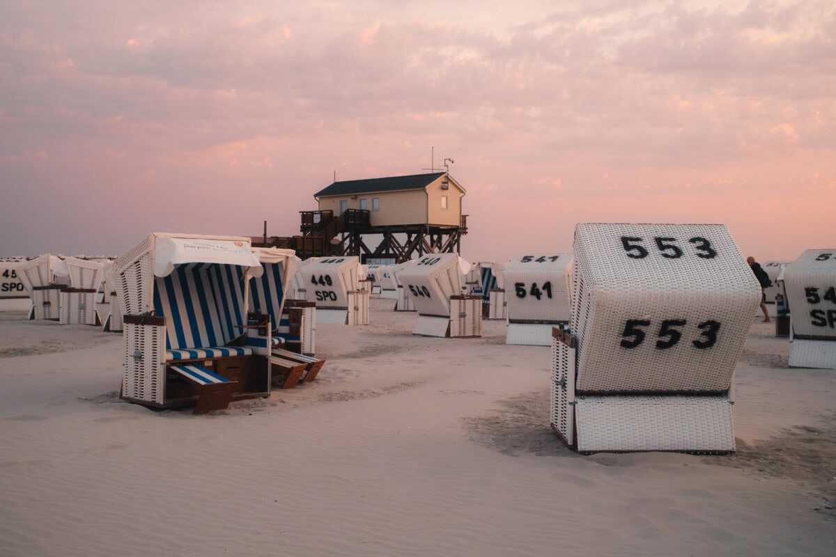 Zahlreiche weiße Strandkörbe am Sandstrand von St. Peter-Ording in der Abenddämmerung, mit einem Pfahlbau im Hintergrund.
