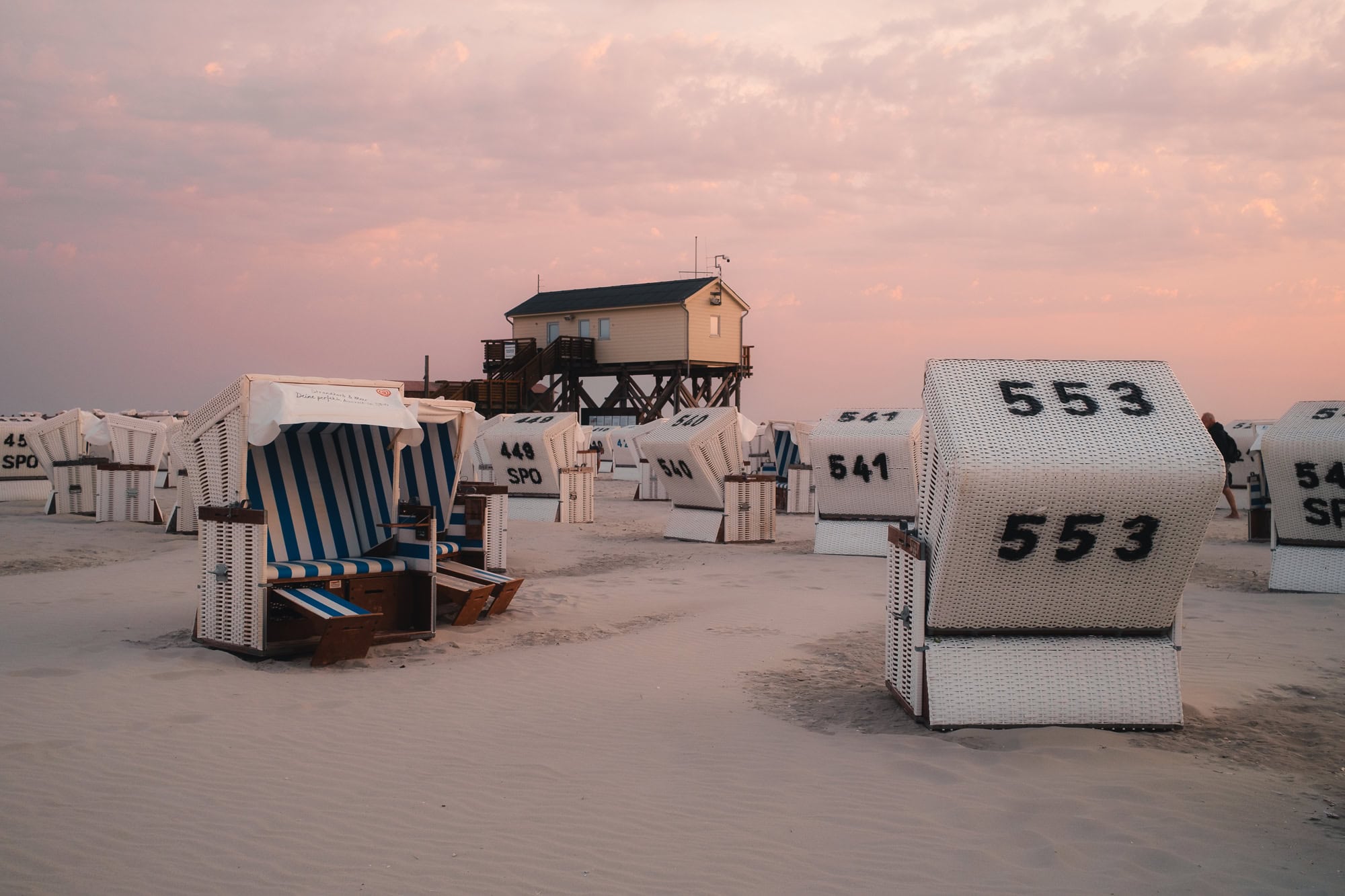 Zahlreiche weiße Strandkörbe am Sandstrand von St. Peter-Ording in der Abenddämmerung, mit einem Pfahlbau im Hintergrund.