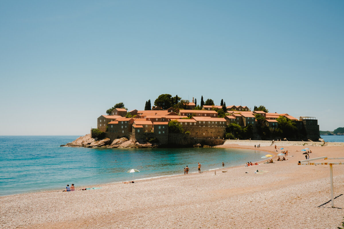 Blick auf Sveti Stefan in Montenegro vom öffentlichen Kiesstrand aus, mit Badenden und Sonnenschirmen im Vordergrund.